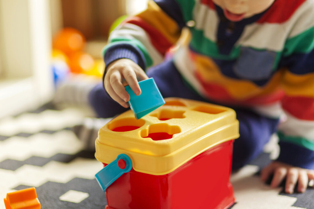 toddler learning shapes with a yellow and red shape sorter, placing a blue square block into the matching slot on the lid