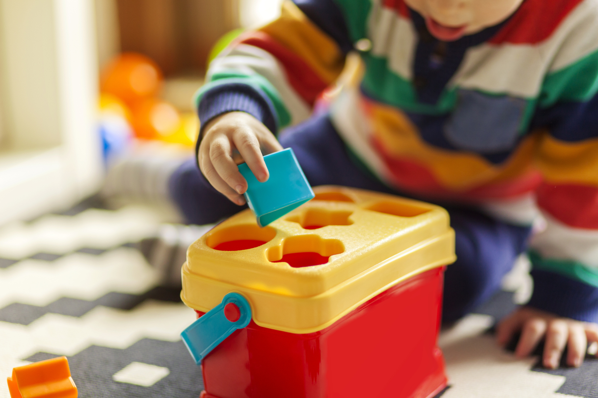 toddler learning shapes with a yellow and red shape sorter, placing a blue square block into the matching slot on the lid