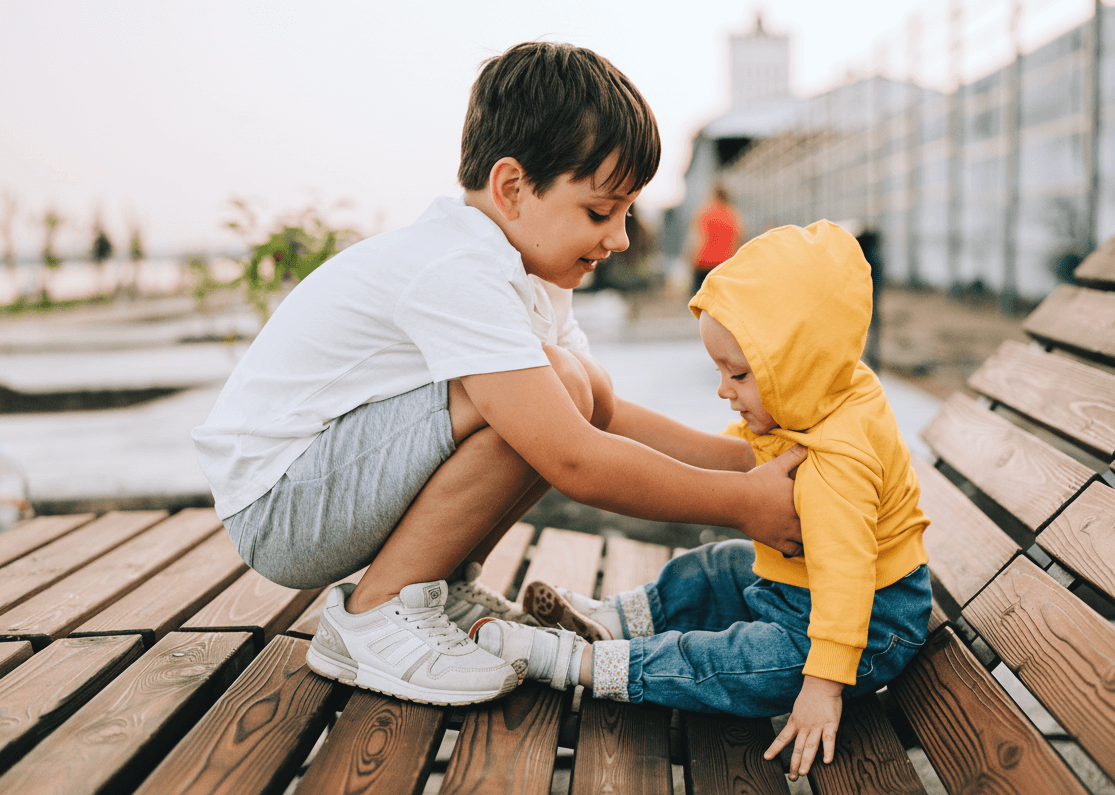 Older child comforting younger sibling outdoors on a bench, fostering empathy and kindness.
