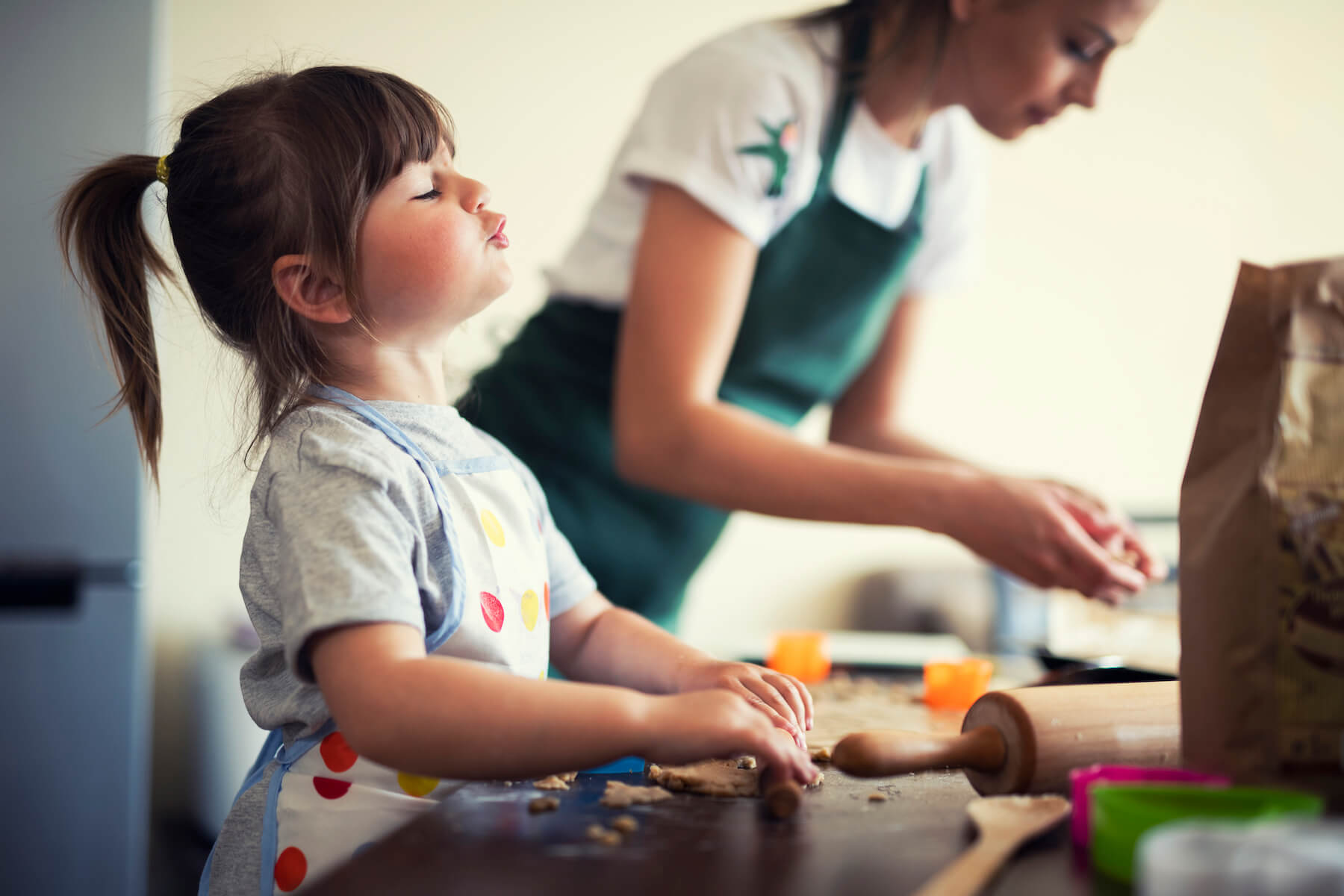 Young child wearing an apron rolls dough on a kitchen counter while an adult beside her prepares ingredients, highlighting hands-on cooking and learning time together.