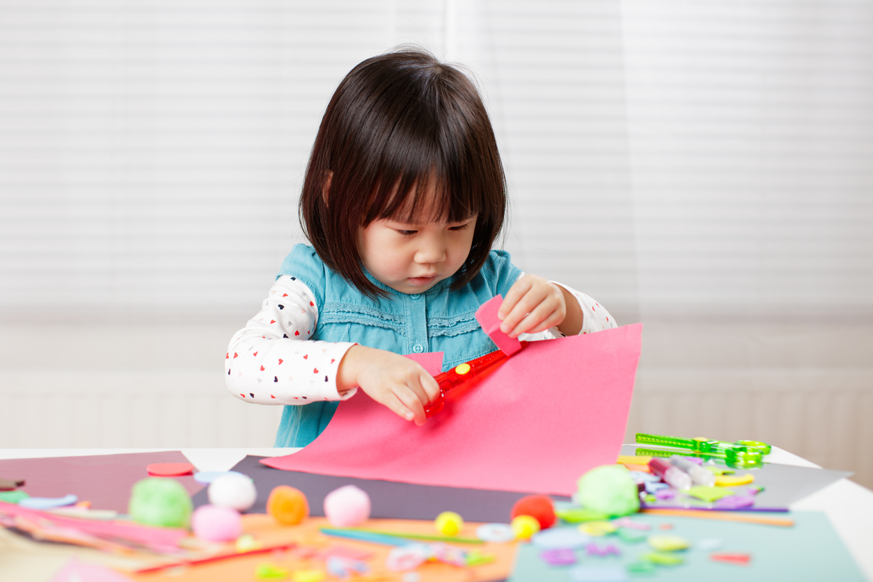Preschool girl using child-safe scissors to cut bright paper during a craft activity at a table.