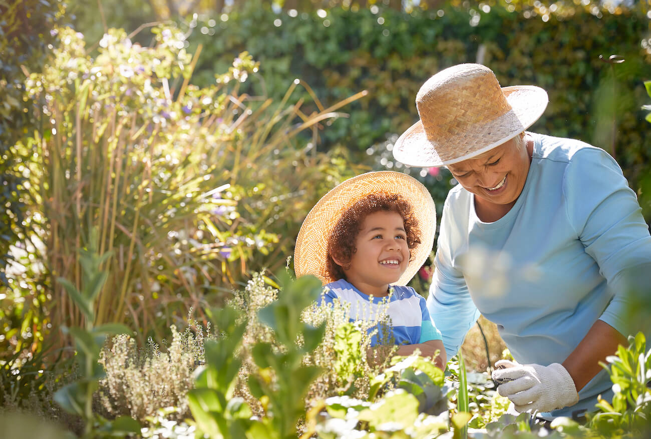 Grandmother and young child wearing sun hats gardening together in a lush backyard, exploring plants and nature as part of everyday learning at home.