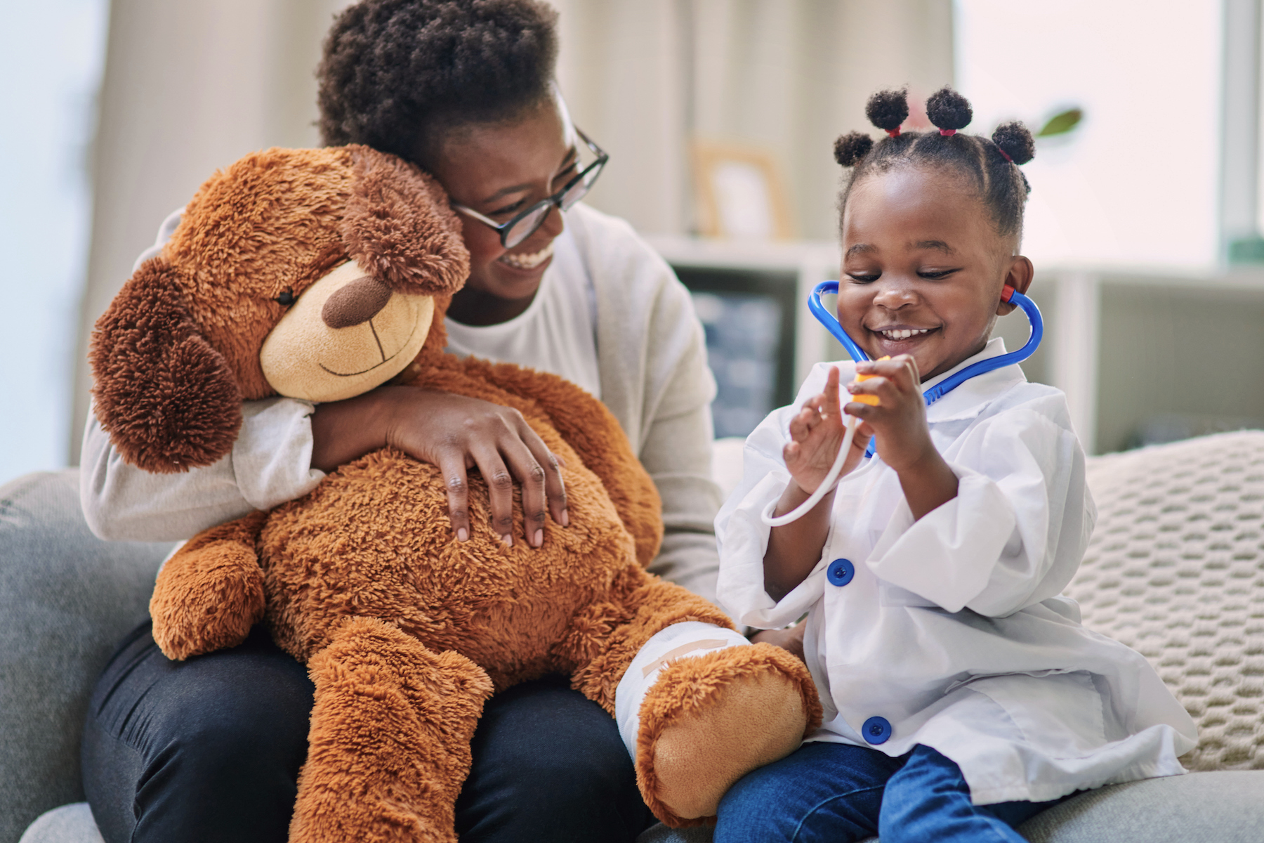 Parent sitting on a couch holding a large teddy bear while a young child in a doctor costume uses a toy stethoscope.