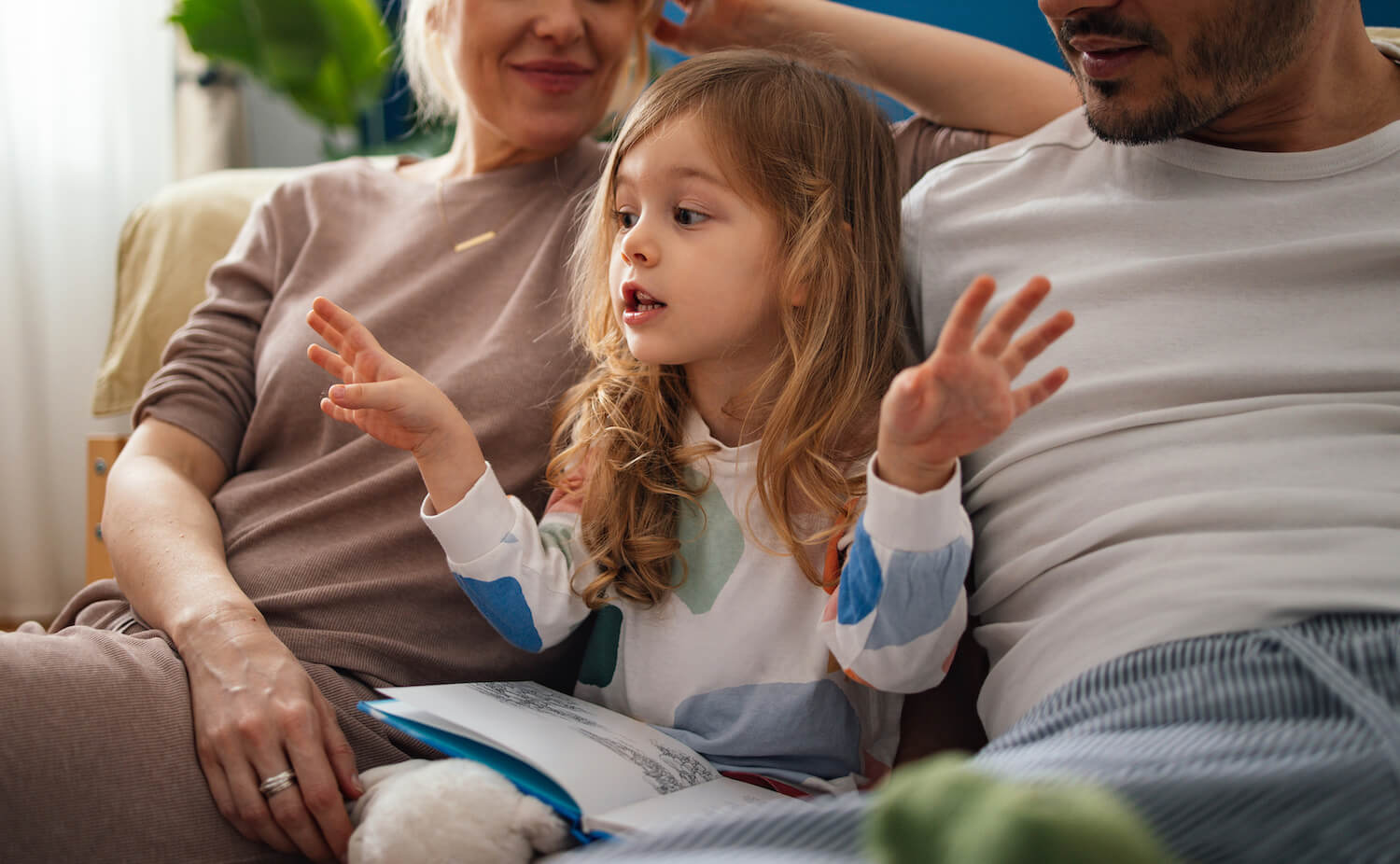 Parents cuddling on the sofa while their young child talks and gestures during shared reading time.