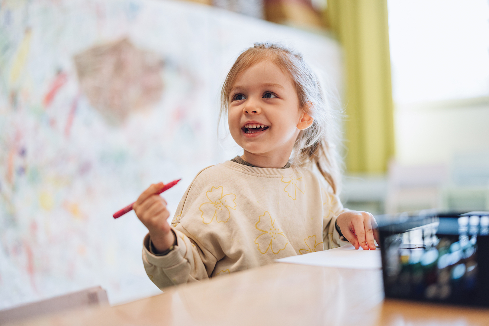 Happy kindergarten girl smiling while drawing with a red crayon at her classroom table, building independence skills.