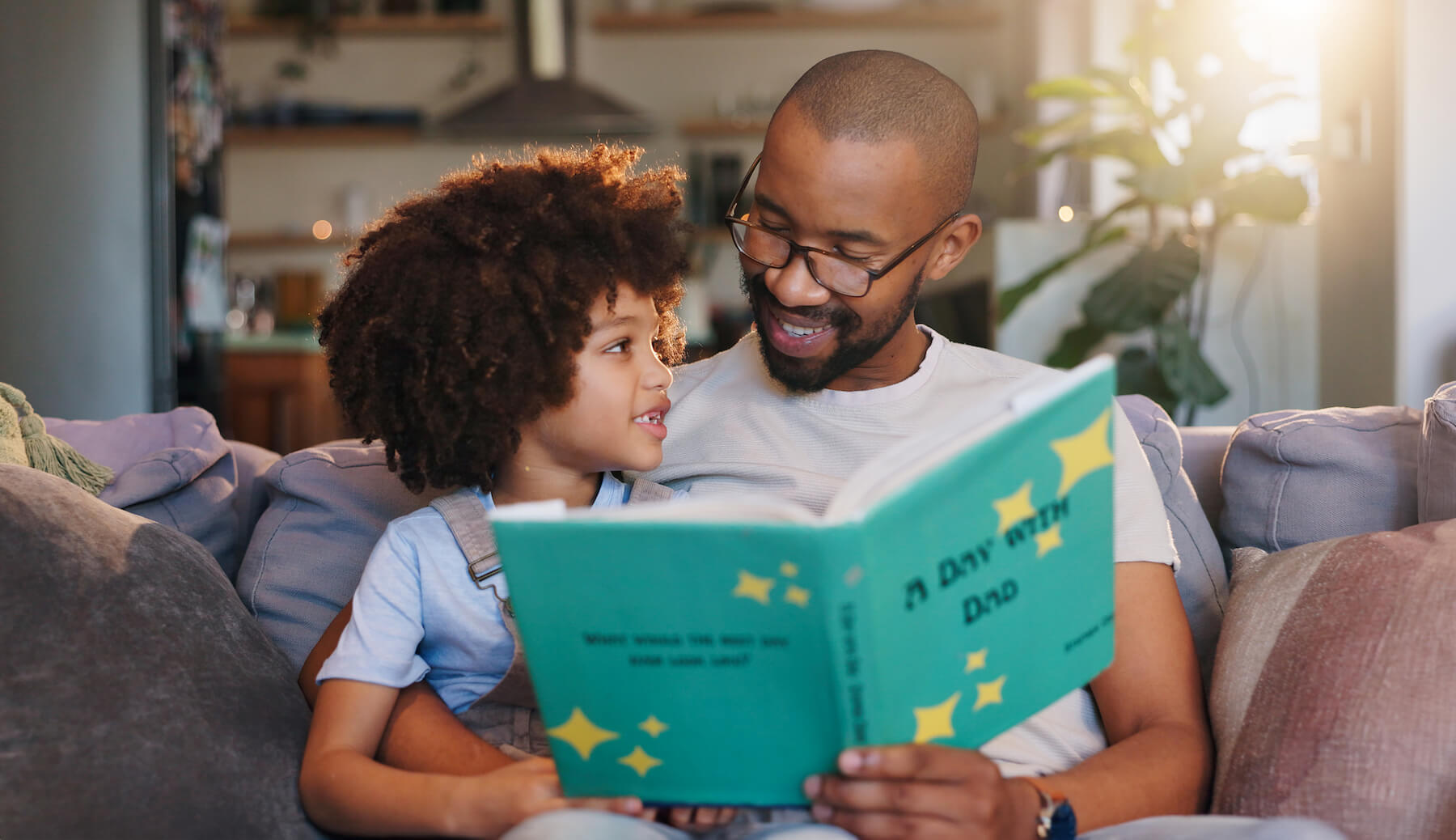 Father reading a bright picture book with his preschooler on a cozy couch in a sunlit living room.
