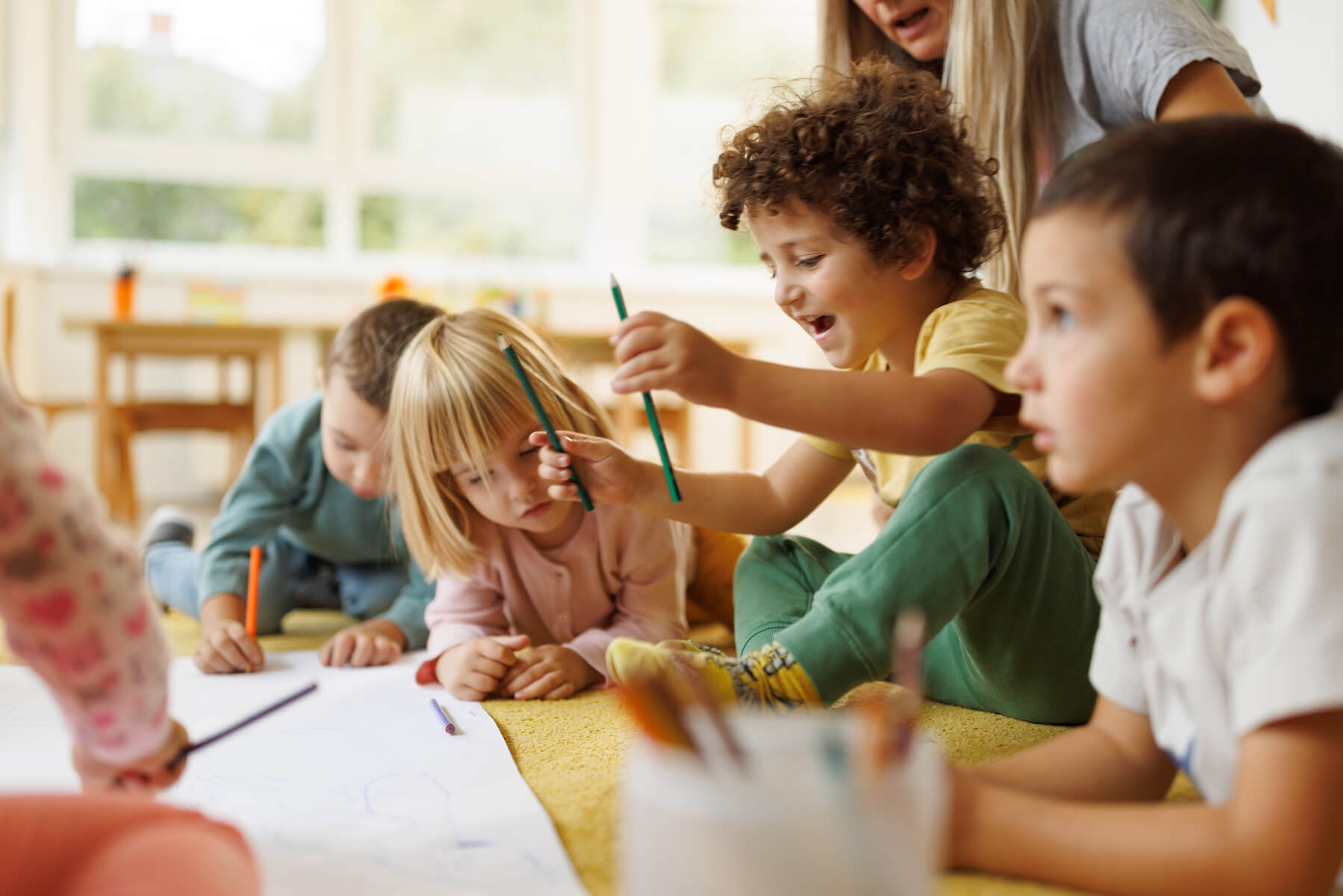 Children sitting together on the floor, drawing and sharing colored pencils, supervised by a teacher in a bright classroom.