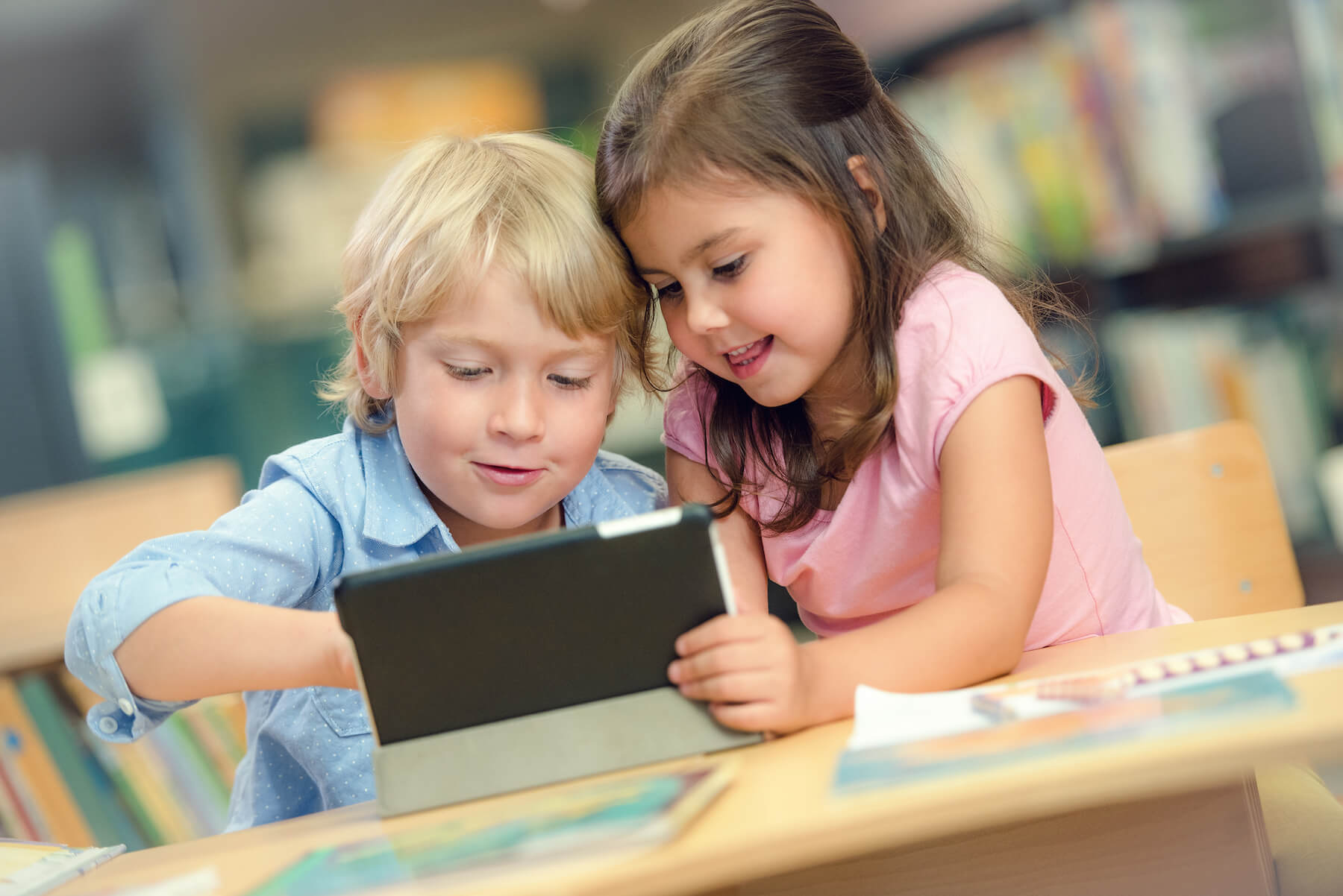 Two young children smiling and using a tablet together at a classroom table.