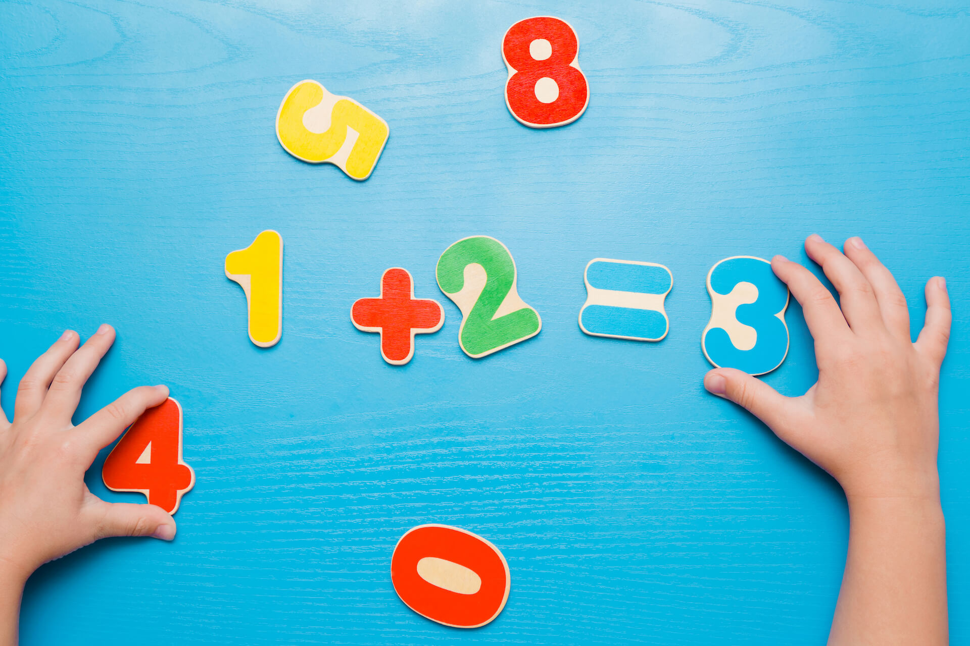 Child’s hands arranging colorful number and math symbol magnets on a blue table to show 1 + 2 = 3, with extra numbers nearby