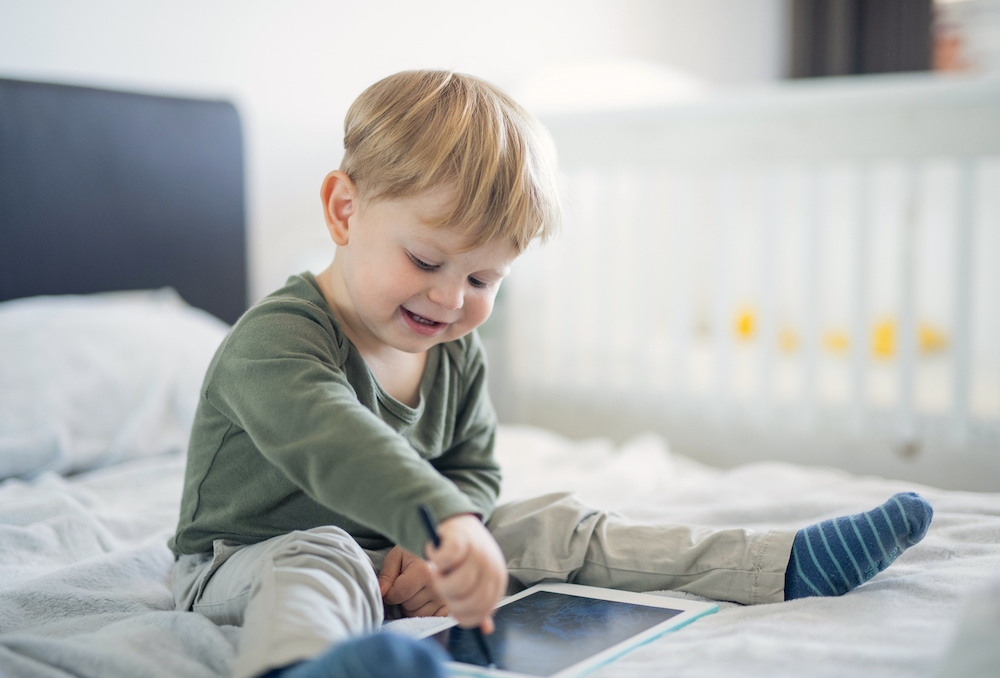 Young child sitting on a bed using a tablet with a stylus, in a softly lit bedroom illustrating the need to choose ad‑free, age‑appropriate, and privacy‑safe apps for young children