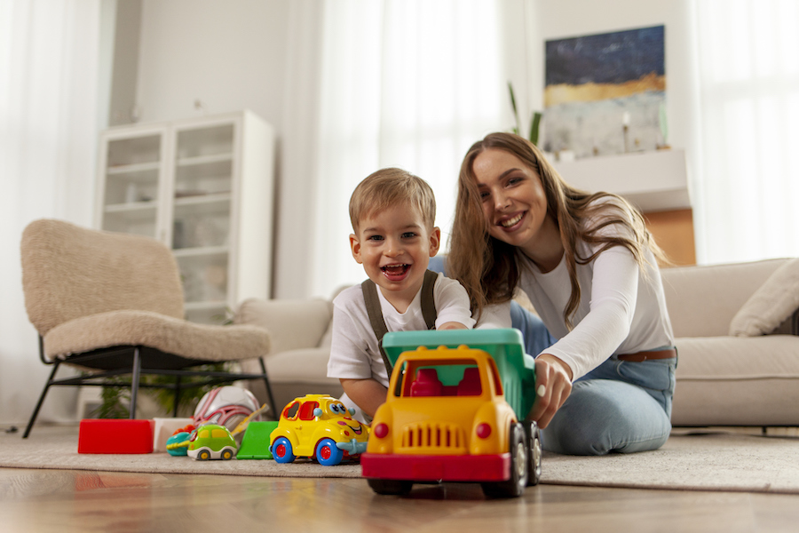 Joyful preschool boy and mom play with colorful toy dump truck on living room floor, boosting fine motor skills.
