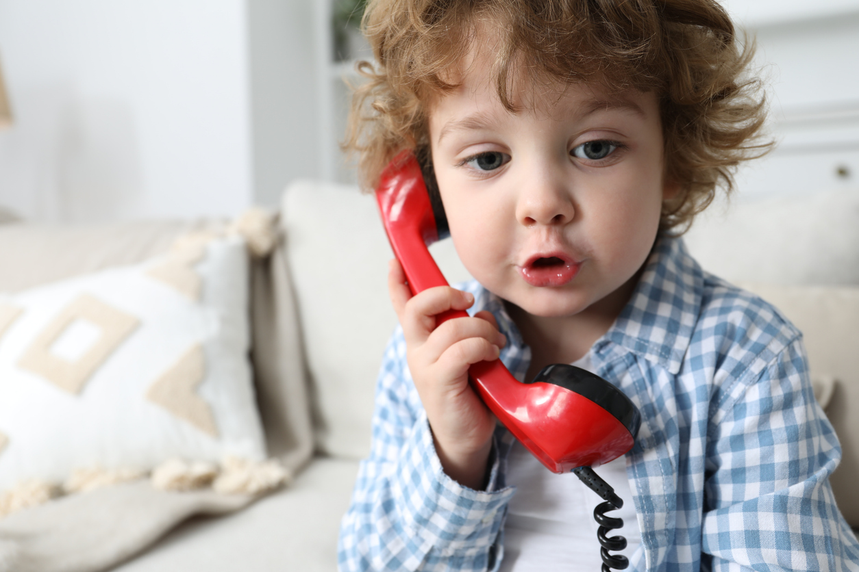 Toddler in a blue checkered shirt holds a red toy phone to their ear while sitting on a beige couch.