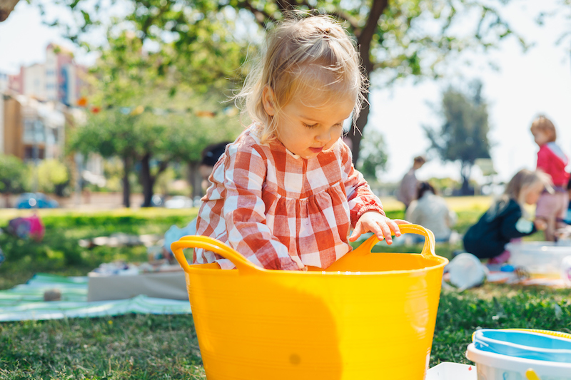 A 2-year-old child sitting on grass outdoors, exploring sensory play by scooping water with toys from a bright yellow tub.