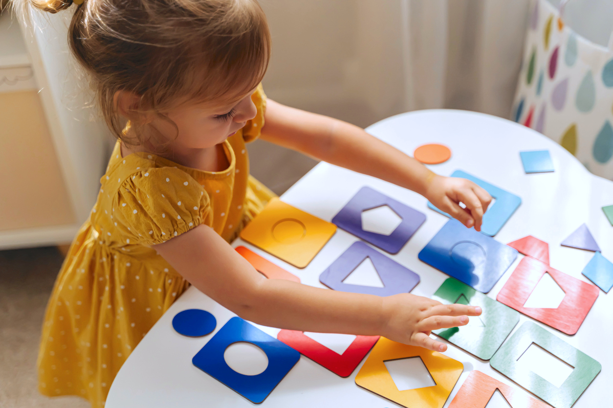 Toddler girl plays with a wooden shape sorter on a table, matching colors and shapes.