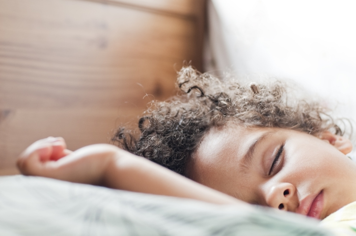 Young child with curly hair sleeps peacefully sideways in wooden crib, arm over edge, on green sheet.