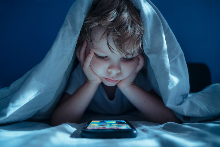Toddler hiding under blanket using phone at bedtime