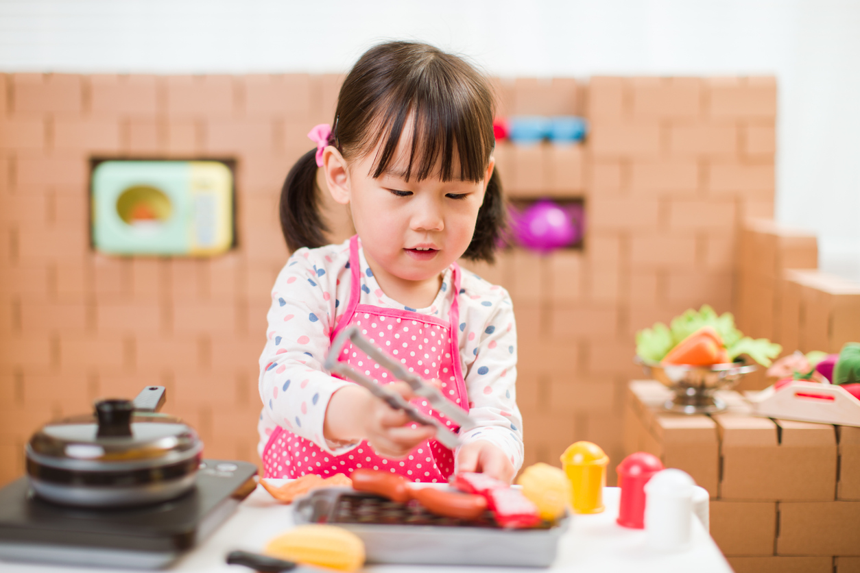 Preschool girl uses kitchen tongs in a pretend play kitchen, practicing fine motor control and grip strength.