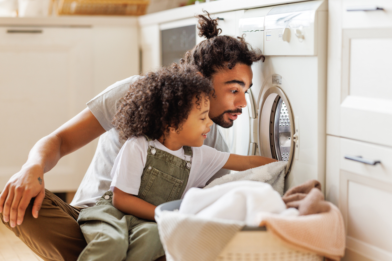 Father and son loading laundry into a front-loading washing machine at home.