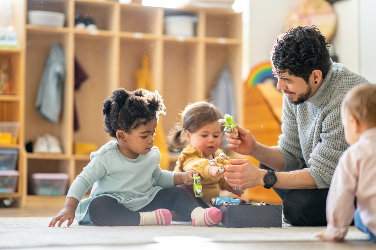 Early childhood teacher sits on the floor with toddlers, handing out instruments from a box to practice sharing and taking turns.