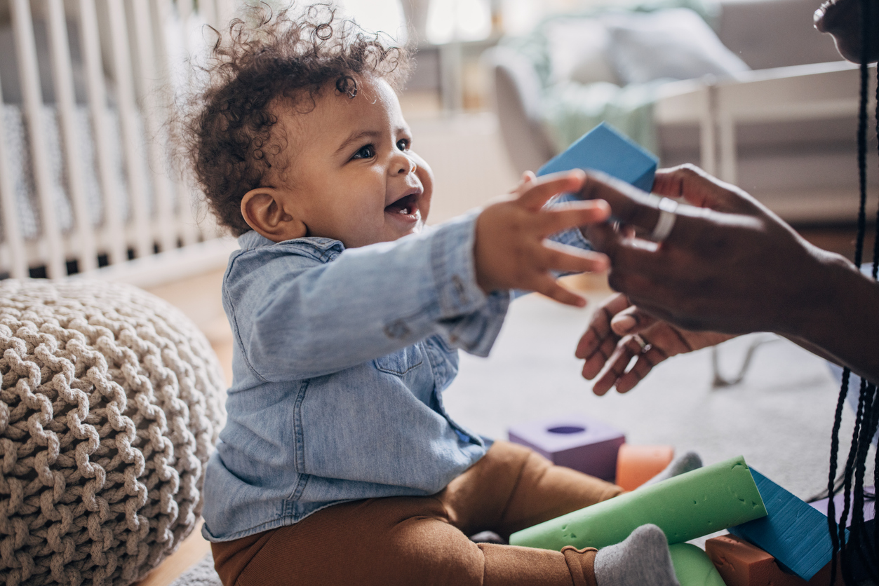 Smiling toddler reaches for a blue block offered by a caregiver while playing with colorful toys on the floor.