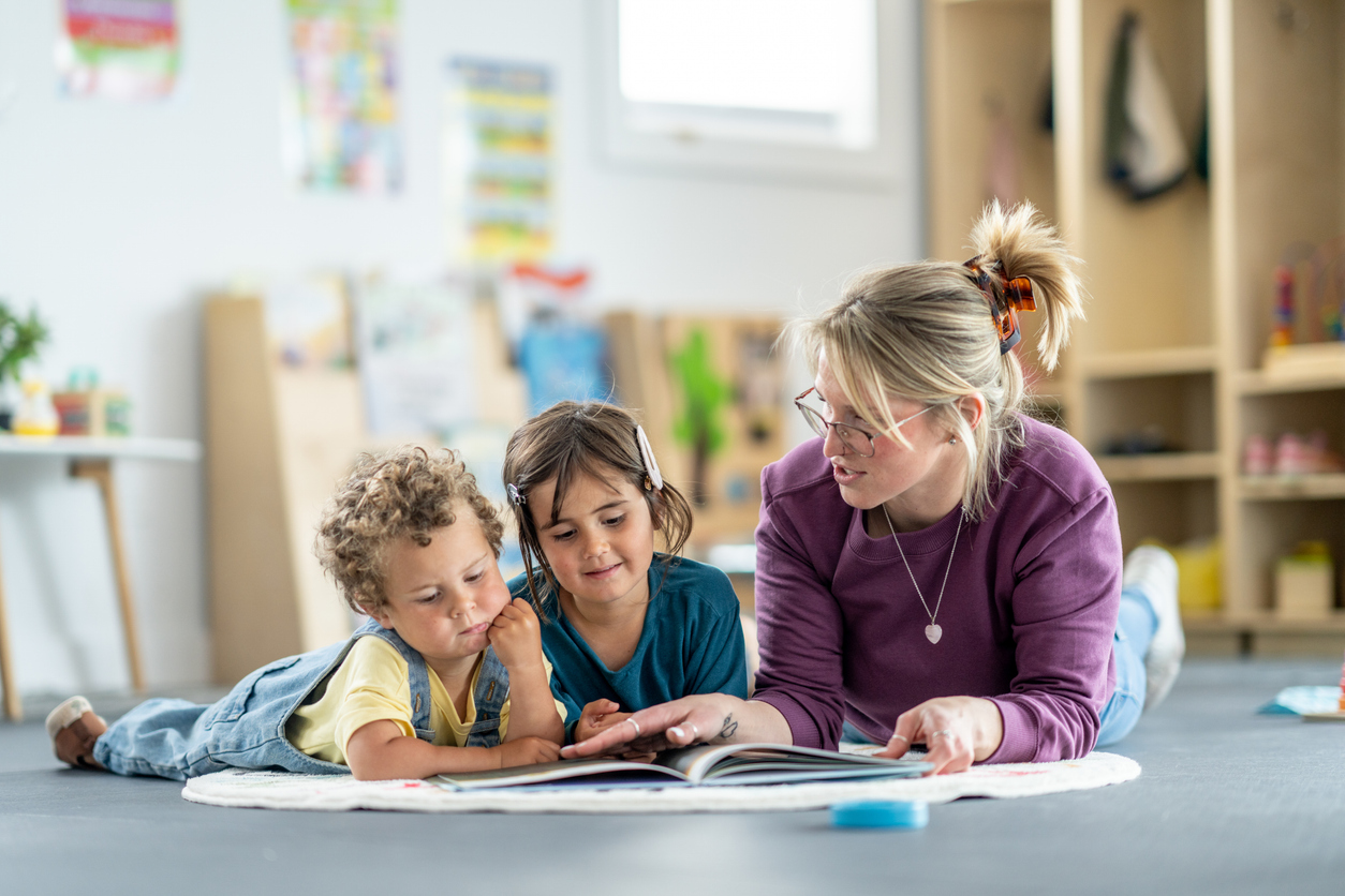 Preschool teacher reads an alphabet picture book with two young children on a rug in a classroom.