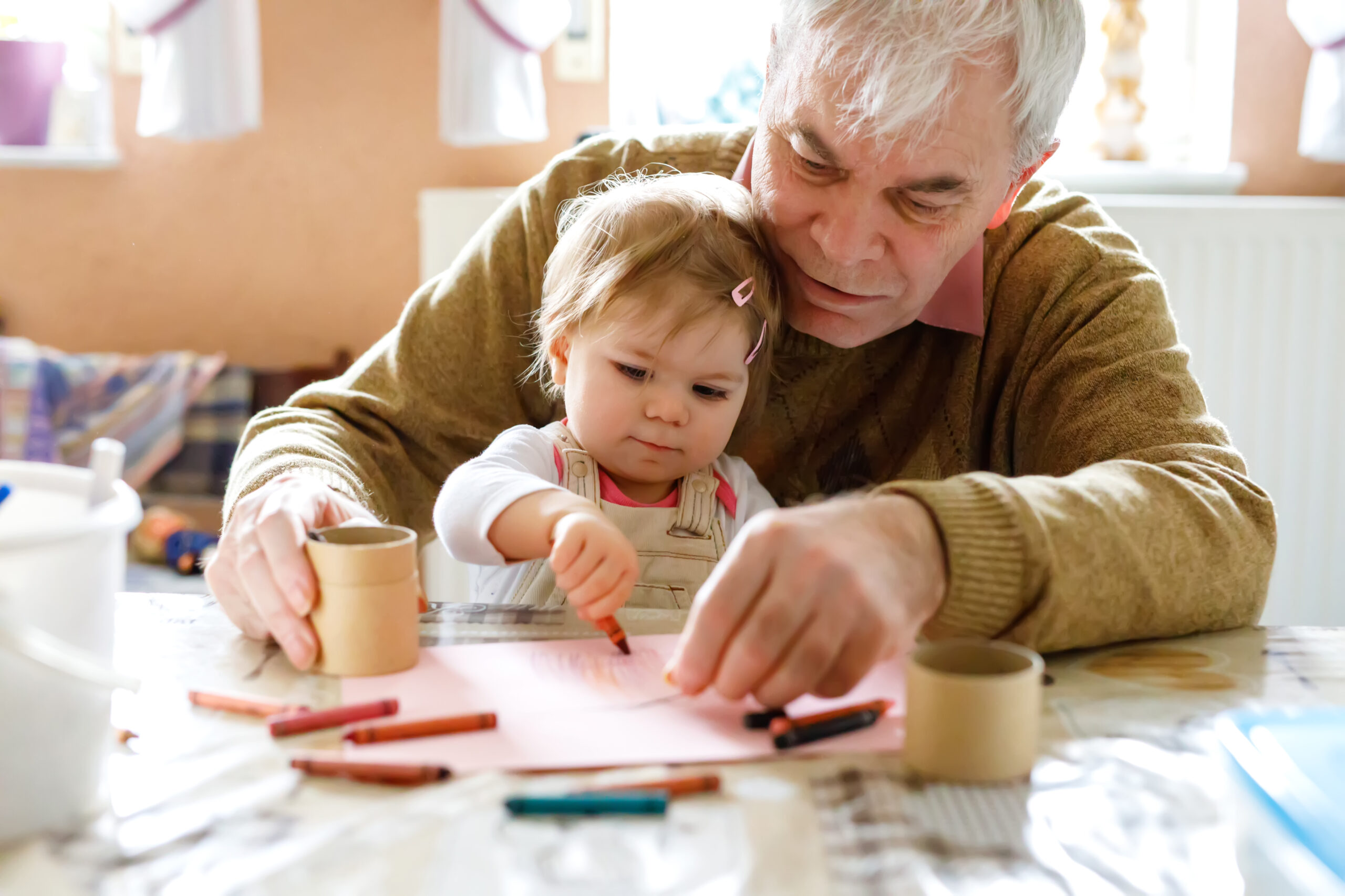 Grandfather helping toddler draw with crayons at home, supporting early learning and bonding