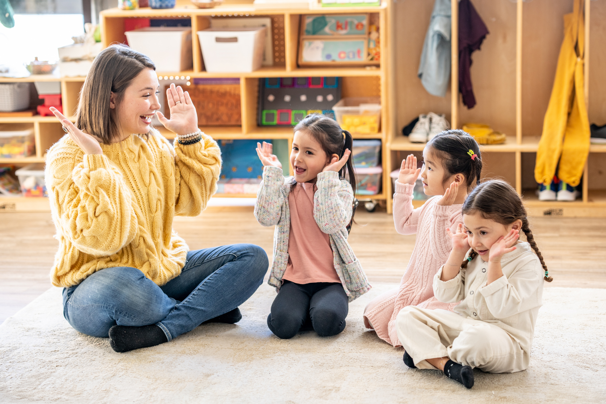 Preschool teacher and three young girls sitting on a classroom floor doing hand gestures together during a nursery rhyme activity